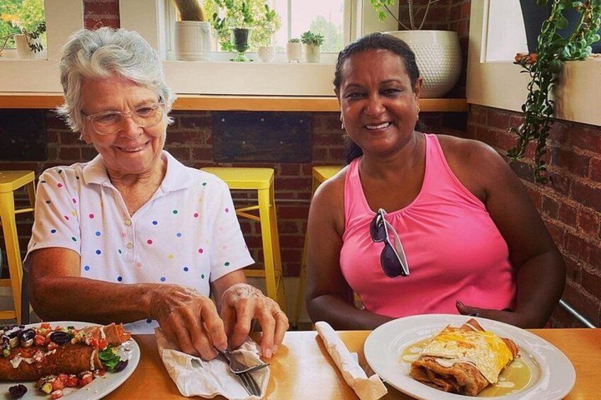 Two people sitting at a table with food, one wearing a polka-dotted shirt and the other in pink, in a cafe with brick walls and plants. 5