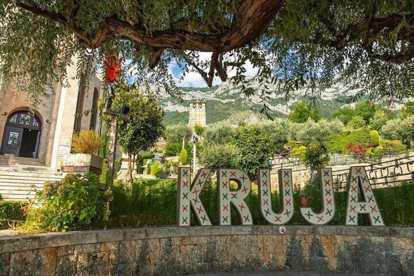 Een stenen muur met de tekst 'KRUJA' in grote letters, een kerk met een donkere deur, en een bergachtig landschap op de achtergrond. 4
