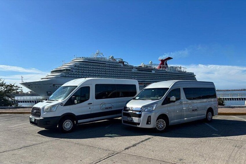 Two white vans parked on a concrete surface with a large cruise ship in the background. 1
