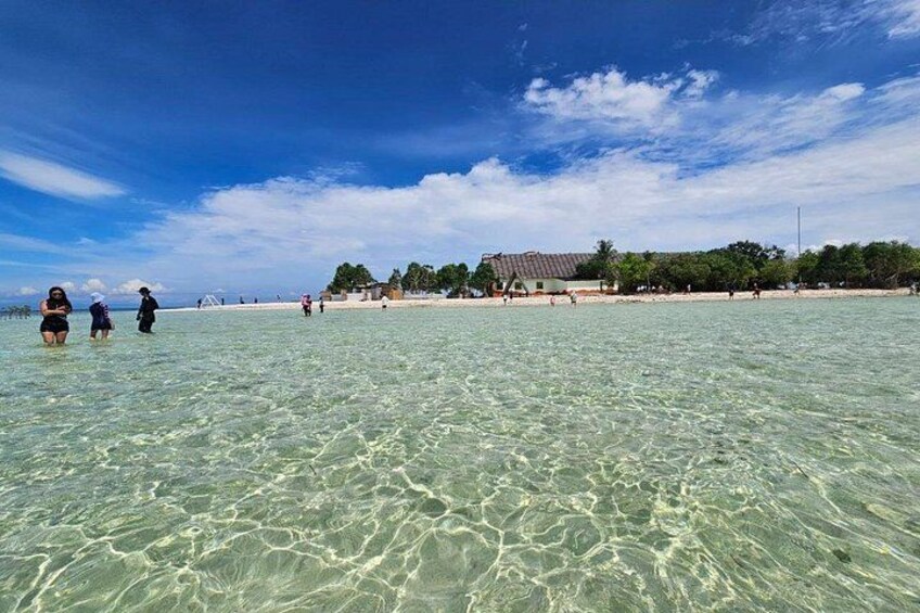 A beach with clear shallow water, people walking, and a building with a tiled roof in the background. 2