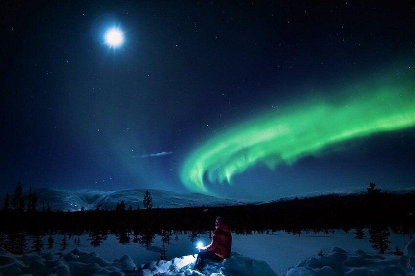 A person in a red jacket sitting on snow with the Northern Lights in the background. 1
