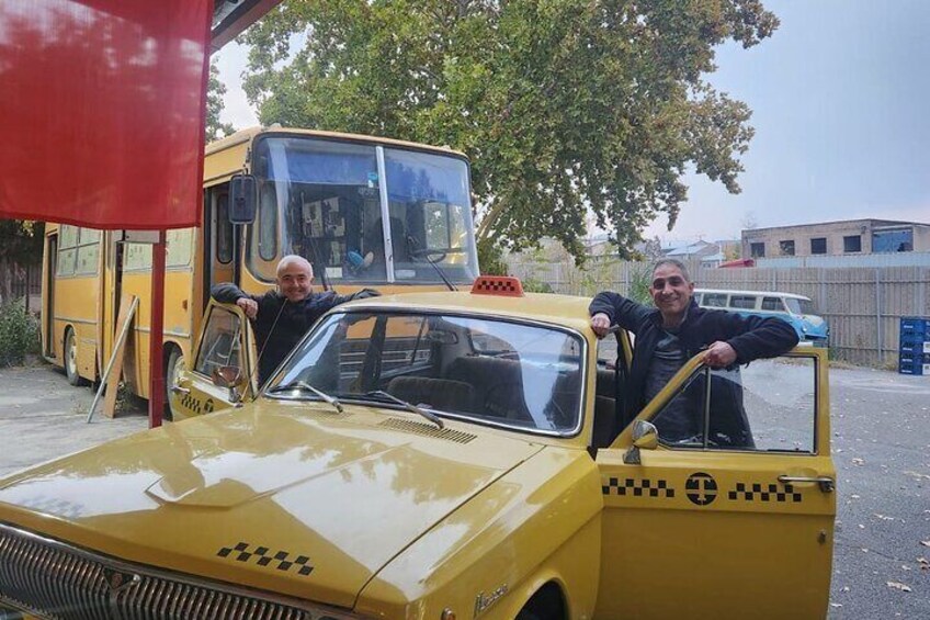 A yellow taxi with two people leaning out of the door, parked in front of a bus. 4