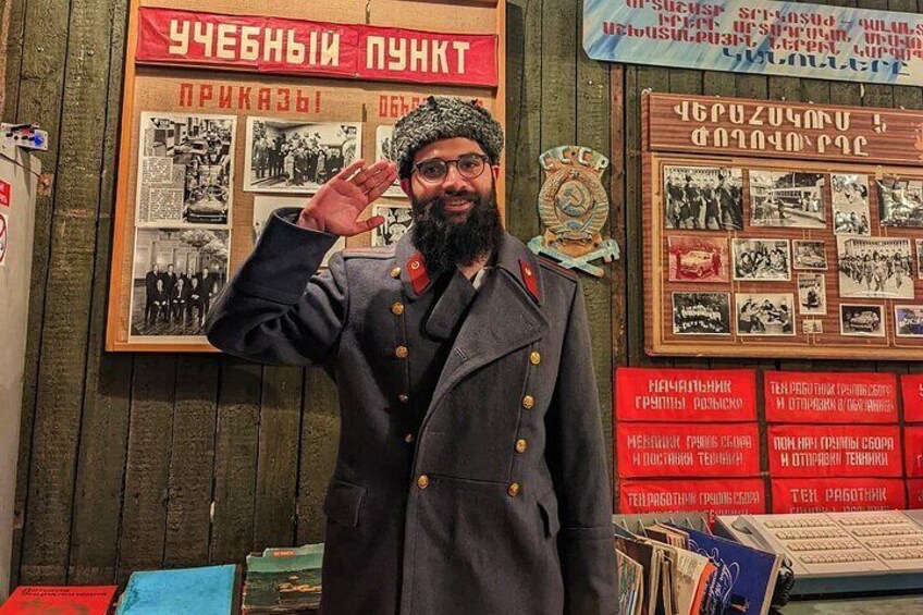 A person in a military uniform saluting. There are books and posters on the wall behind. 1