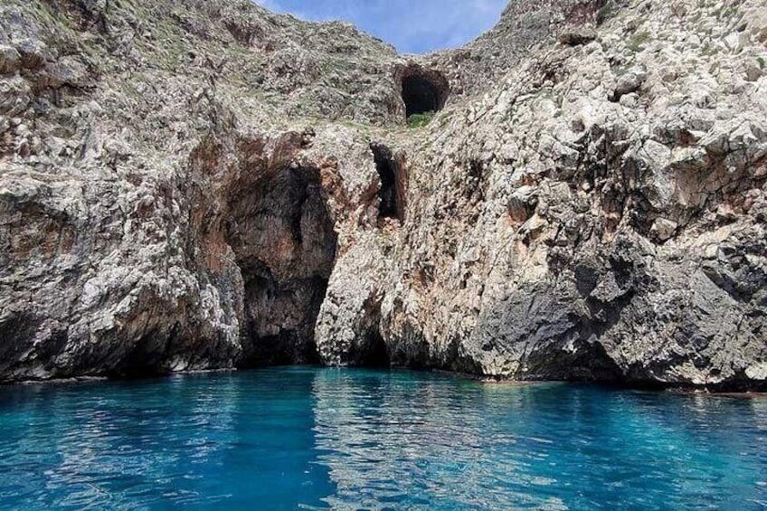 Un paysage côtier rocheux, avec une mer bleue et limpide, l’entrée d’une grotte et un terrain accidenté. 2