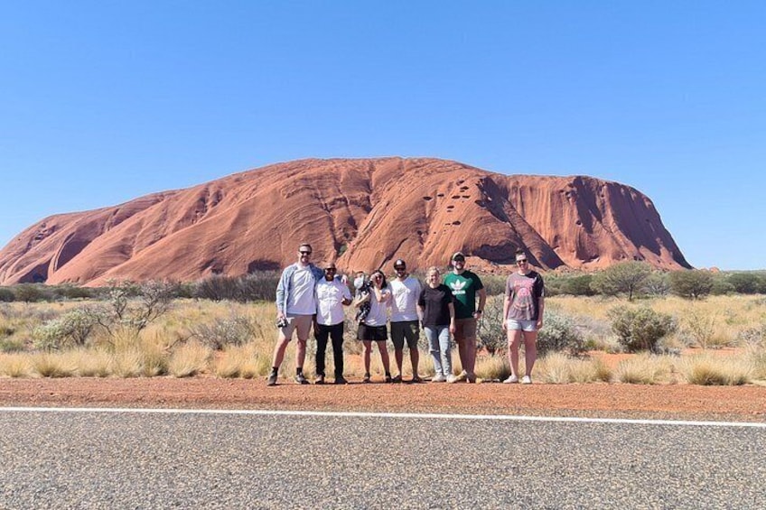 Un groupe de personnes se tient sur une route, avec une grande formation rocheuse rouge en arrière-plan. 3