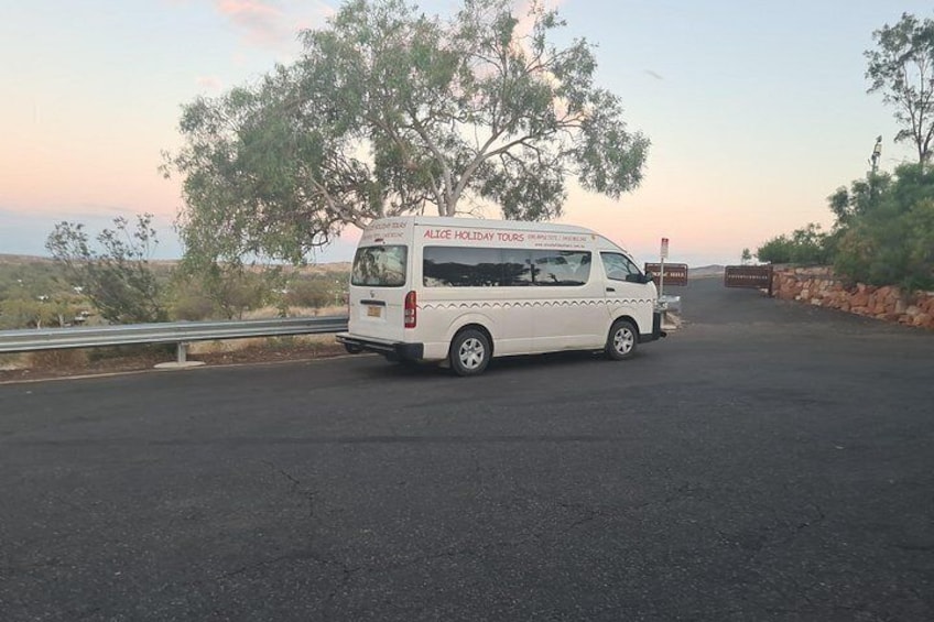 Un fourgon blanc, sur lequel on pouvait lire « ALICE HOLIDAY TOURS », était garé sur une route bordée d’une glissière de sécurité, d’arbres, et le ciel bleu se dégageait à l’arrière-plan. 1