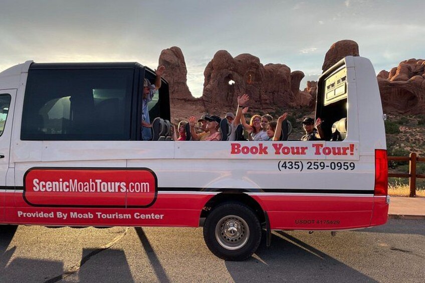 A white tour bus with 'Book Your Tour!' and a phone number on the side, parked in a desert with rock formations in the background. 5