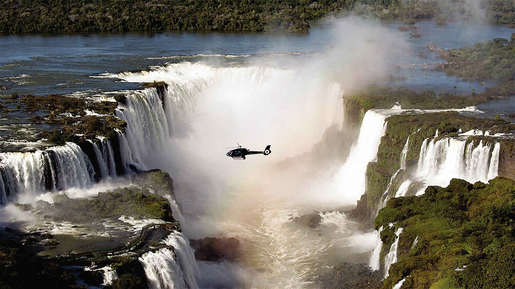 A helicopter flying over a large waterfall surrounded by lush greenery. 1