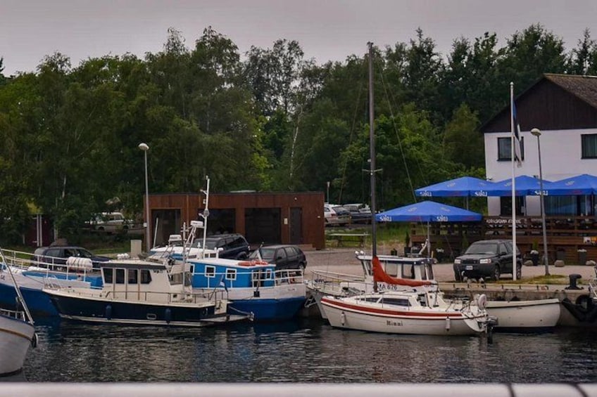 Een jachthaven met verschillende boten die aangemeerd liggen, een gebouw met blauwe parasols en een bebost gebied op de achtergrond. 4