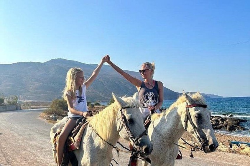 Two people riding horses on a road with a mountainous landscape and a body of water in the background. 3