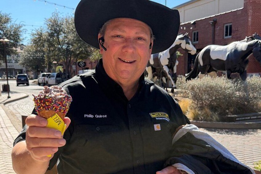 Una persona sosteniendo un donut con envoltorio amarillo, vistiendo una chaqueta negra con un gafete y un sombrero negro. 2