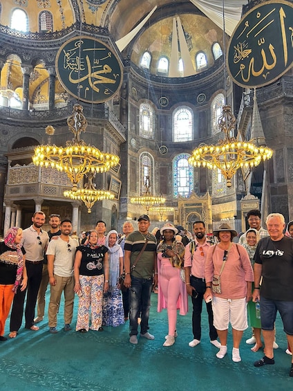Ryhmä ihmisiä historiallisessa rakennuksessa, jossa on koristeellisia kattokruunuja ja seinillä arabialaisia kalligrafiakoristeita. 1