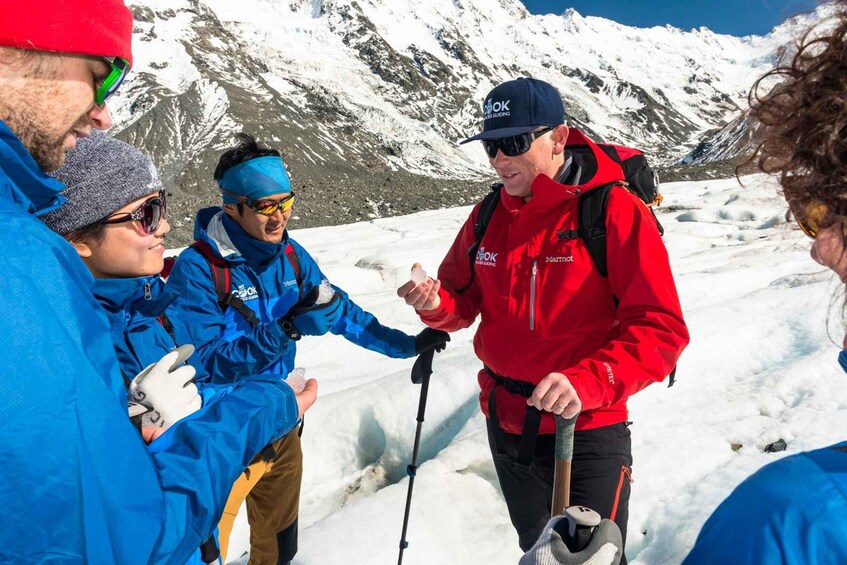 A group of people in a snowy mountainous area, some wearing red jackets and others in blue, with snow-covered peaks in the background. 2