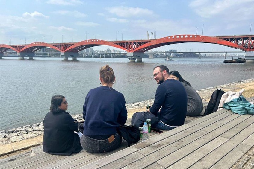 Three people sitting on a wooden dock by a river with a red bridge in the background. 4