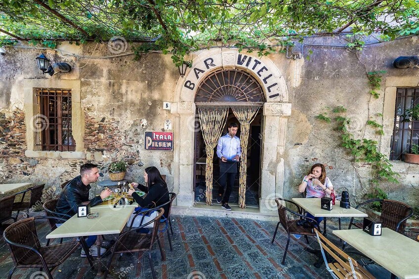 A rustic outdoor cafe with tables and chairs, a man standing at the entrance, and a sign reading "BAR VITELLI". 4