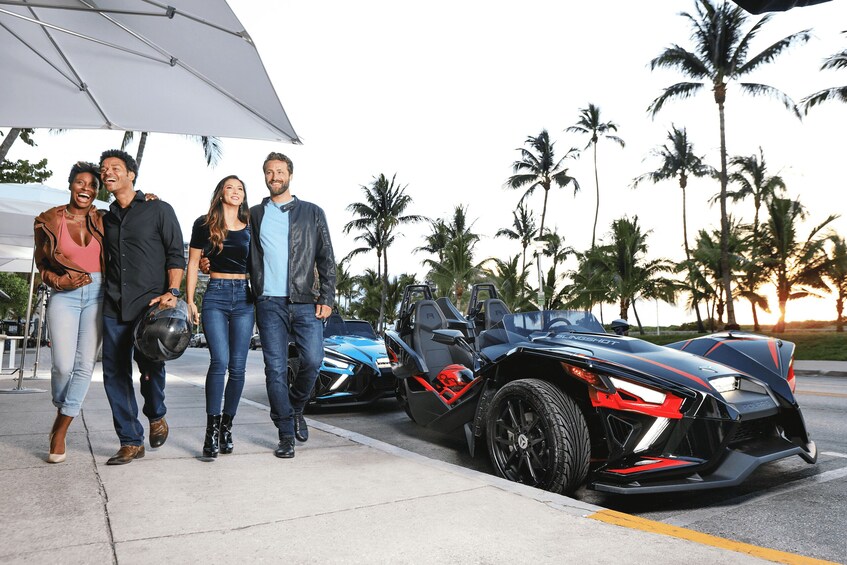A group of people walking past a black sports car and a blue sports car on a sunny day with palm trees in the background. 2