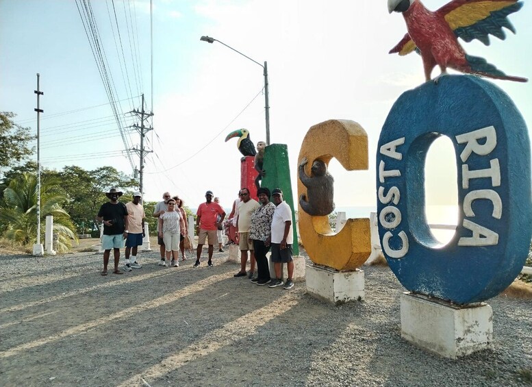 A group of people standing near a large sign with 'Costa Rica' written on it, surrounded by palm trees and power lines. 3