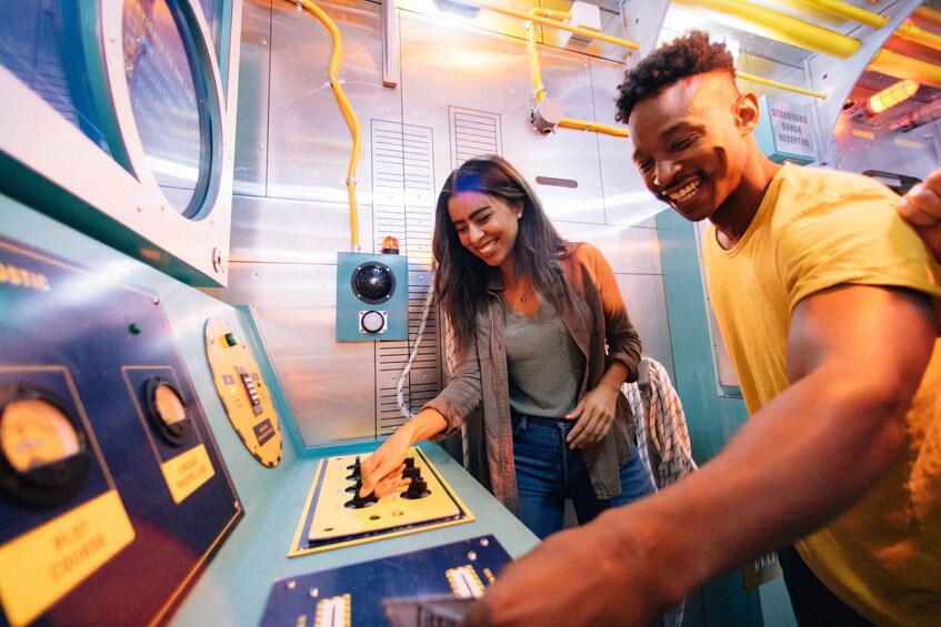Two people interacting with a control panel in a room with industrial elements. 5