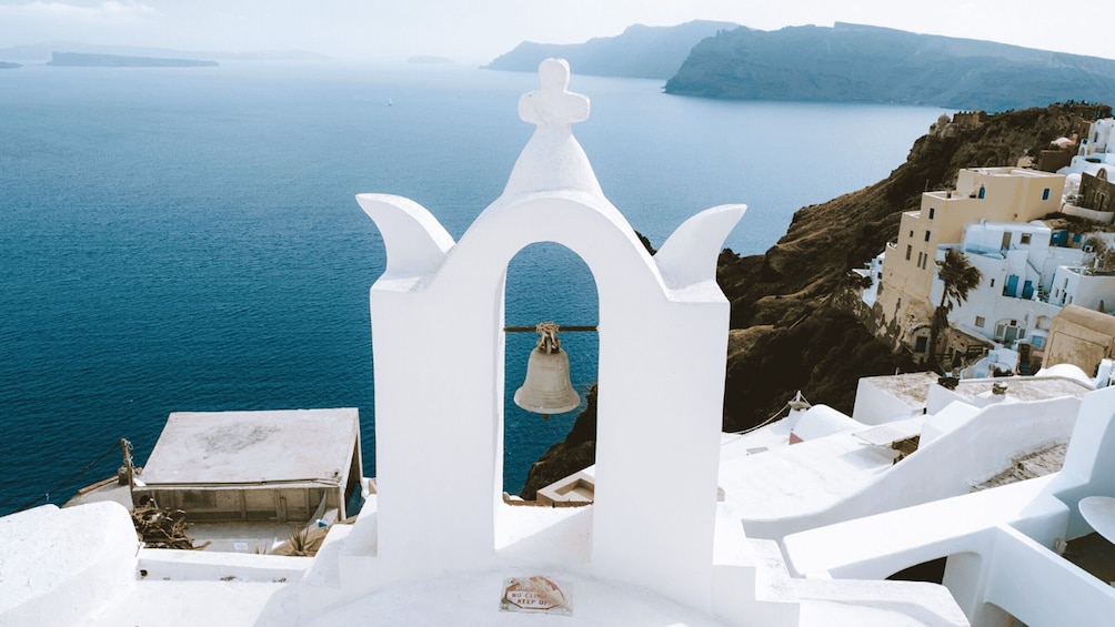 A white bell tower with a bell, overlooking a coastal village with blue waters and white buildings. 5