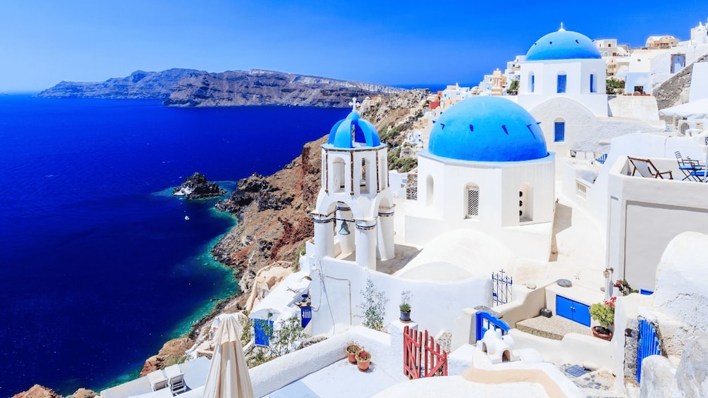 A coastal village with white buildings and blue domes, overlooking the sea and a distant mountain range. 5
