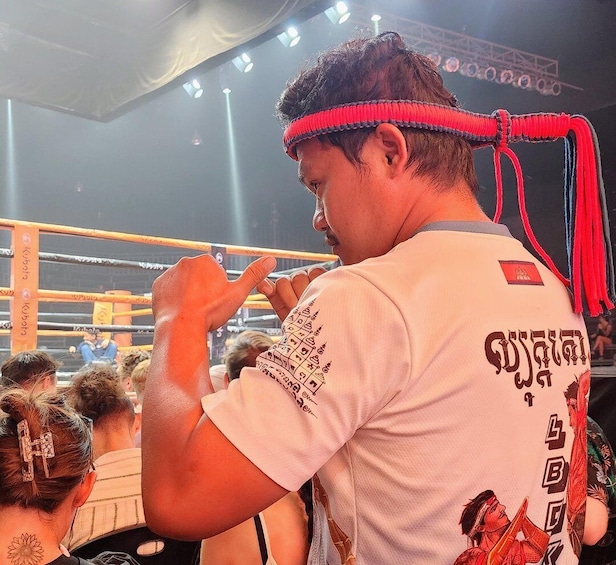 A person in a white shirt with text and graphics, standing in a boxing ring with ropes and a referee in the background. 1