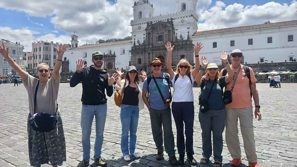 A group of people in a historic plaza with a prominent white building featuring a bell tower in the background. 1
