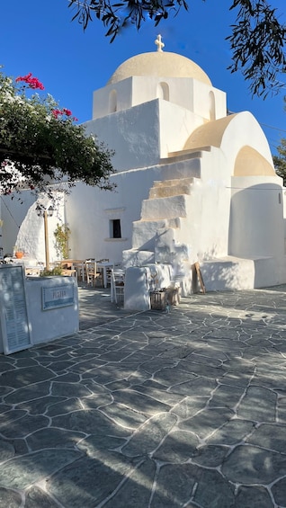 A white church with a dome and a cross on top, surrounded by stone pavement and potted plants. 5
