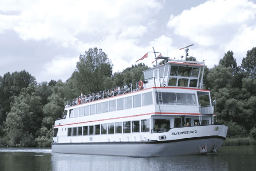 A white riverboat with a red stripe, named 'Zilvermeeuw 2,' sailing on a calm river. 2