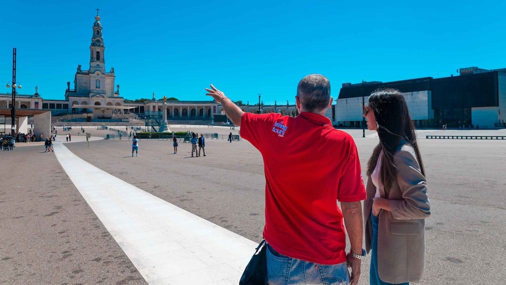 A man pointing towards a large building with a tower, a wide open plaza, and a few people walking. 3