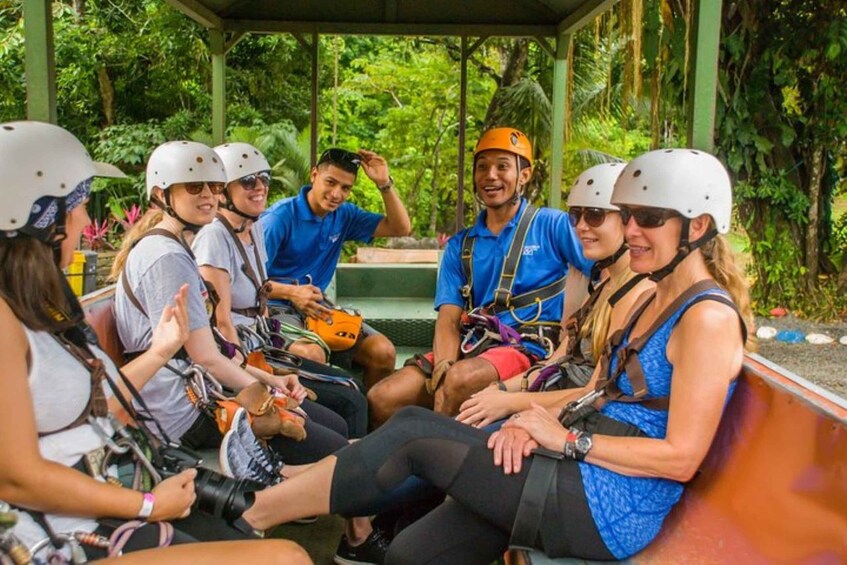 A group of people in helmets and harnesses sitting in a covered area with trees in the background. 4