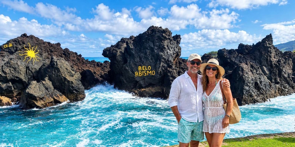 A couple standing by a rocky coastline with waves crashing against the rocks. The text 'BELLO ARISMIO' is visible on one of the rocks. 1
