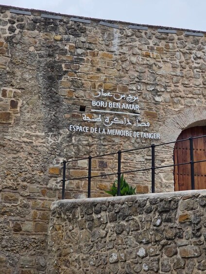 A stone wall with a metal gate and a wooden door. There is a sign with text in multiple languages. 4