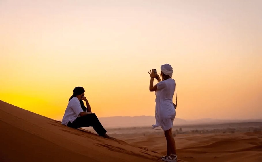 Two people sitting on sand dunes during sunset, one taking a photo, and another holding up a phone. 1