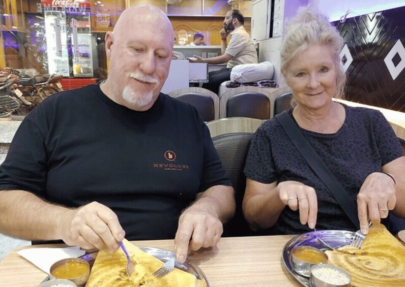 Twee personen zitten aan een tafel met eten, waarvan er een een stuk platbrood snijdt. 5
