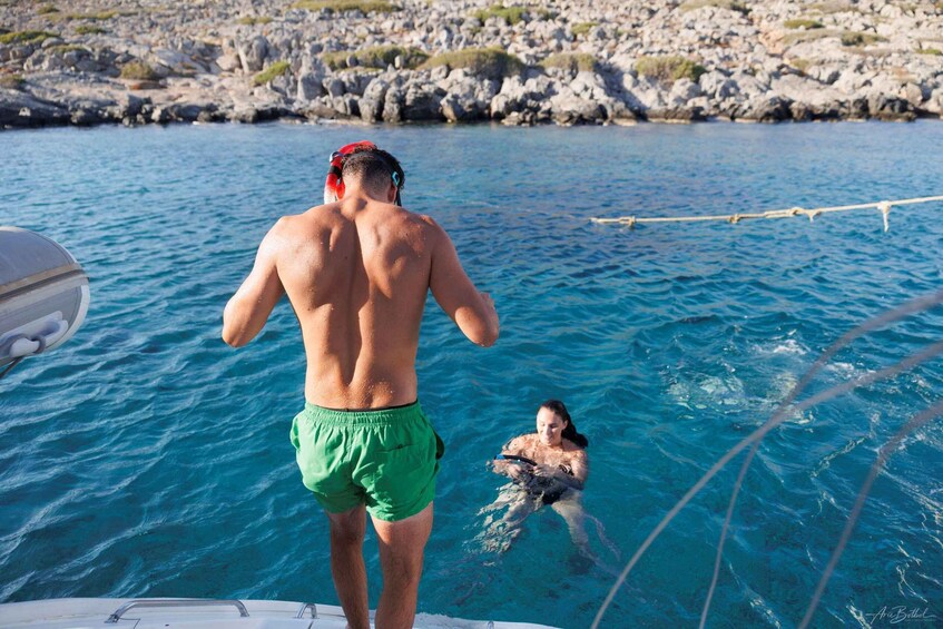 En man i gröna badshorts och en röd badmössa står på en båt och tittar på en kvinna som simmar i vattnet. 2