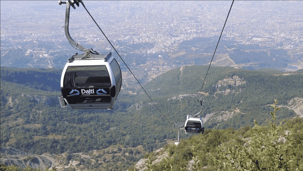 Un teleférico con la marca 'Dajti' suspendido por cables, recorriendo un paisaje montañoso con bosques densos y una ciudad a la distancia. 2