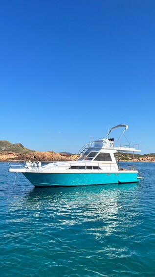 A turquoise boat with a cabin and a canopy, anchored in calm blue waters with a rocky coastline and clear sky in the background. 5