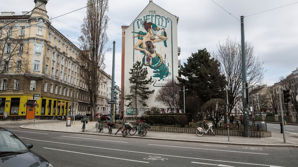 A street with a mural of a woman and birds on a building, bicycles parked along the sidewalk, and a yellow building with a sign. 2