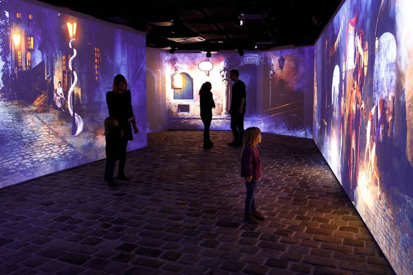Eine Innenansicht einer Ausstellung mit beleuchteten Projektionen an den Wänden, einem Kopfsteinpflasterboden und einigen Besuchern. 2