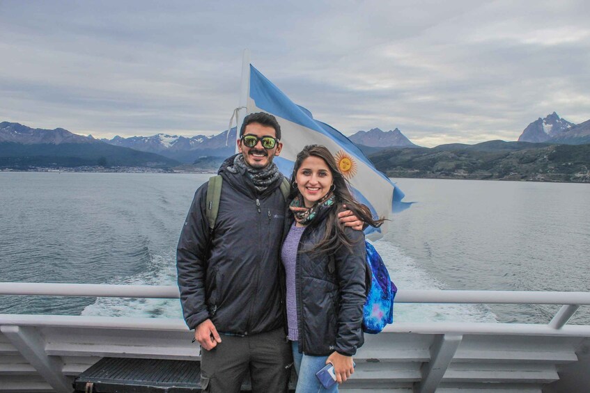 A couple standing on a boat with a flag, mountains in the background, and a lake. 3