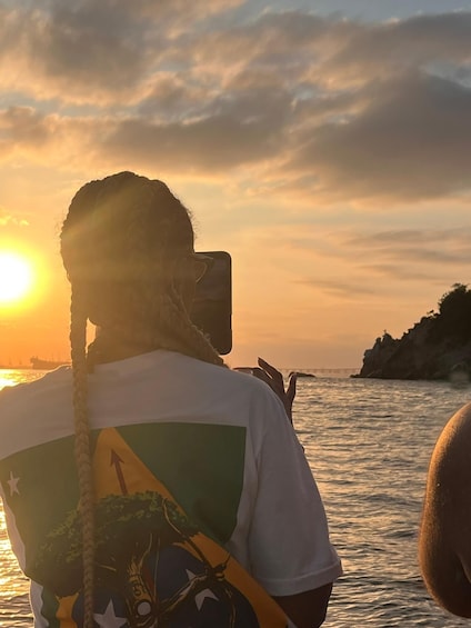 A person with a braided hairstyle, wearing a white t-shirt with a graphic design, standing by the water during sunset. 5
