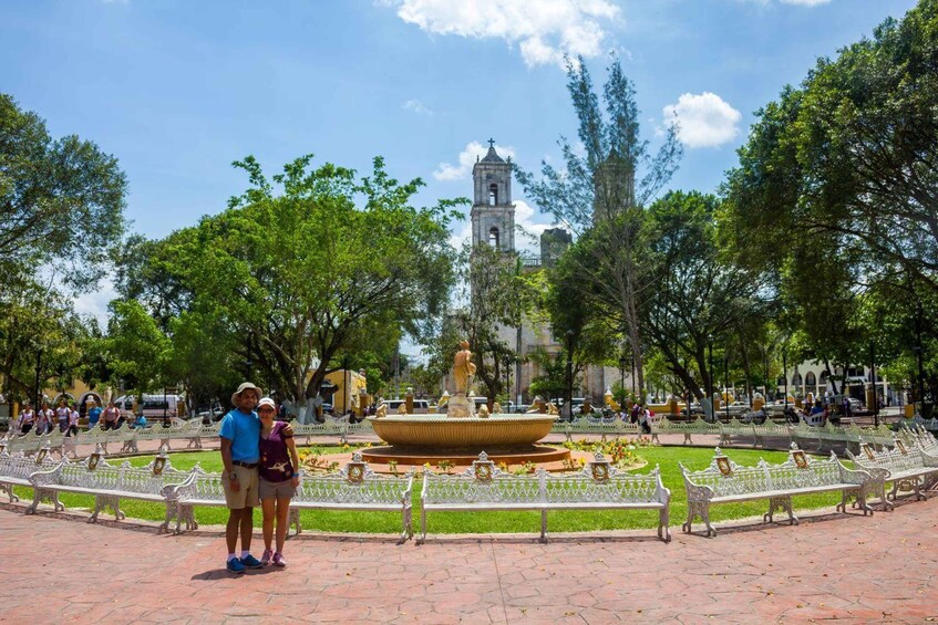A park with a fountain, benches, and a church tower. 3