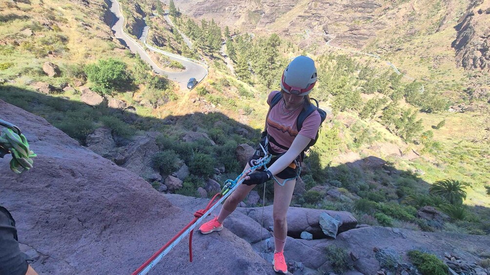 A person climbing a rocky mountain with a helmet and harness, surrounded by a valley with winding roads. 3