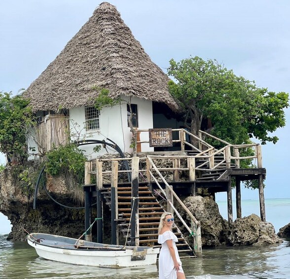 A thatched-roof house on stilts over water, a wooden staircase, and a sign reading "THE ROCK". 1