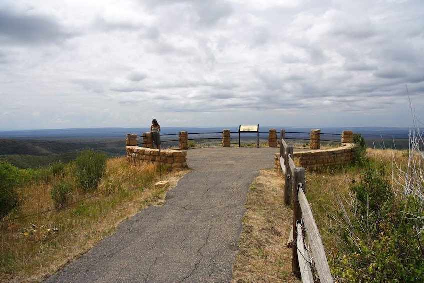 Une route goudronnée mène à une porte en pierre surmontée d’une enseigne, le tout étant entouré d’herbes sèches et de buissons, sous un ciel nuageux. 5