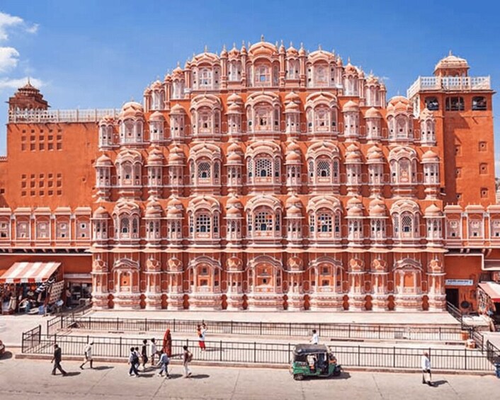 A large, ornate building with multiple domes and arches, surrounded by a paved area with people and a green auto-rickshaw. 1