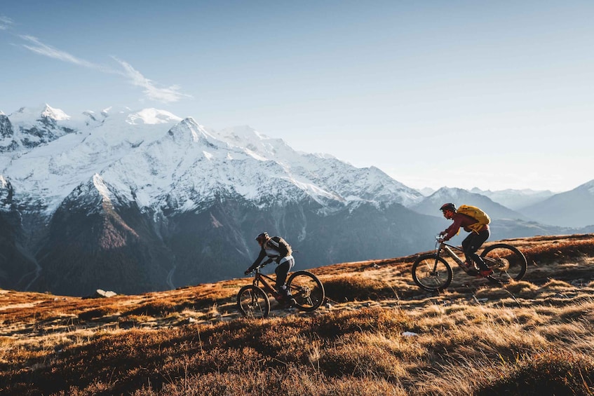 Two cyclists on a grassy mountain trail with snow-capped peaks in the background. 2