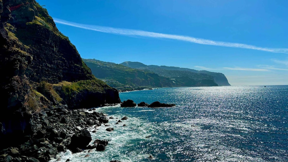 A coastal landscape with rocky cliffs, a clear blue sea, and distant hills. 1