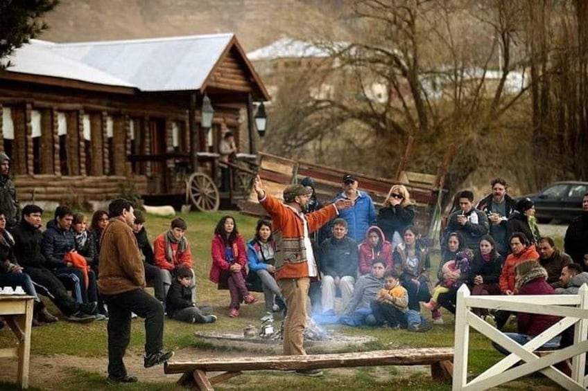 A group of people gathered around a campfire in a rustic outdoor setting with wooden cabins in the background. 1