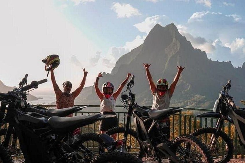 Three people with raised arms, a mountain, and atv bikes. 1